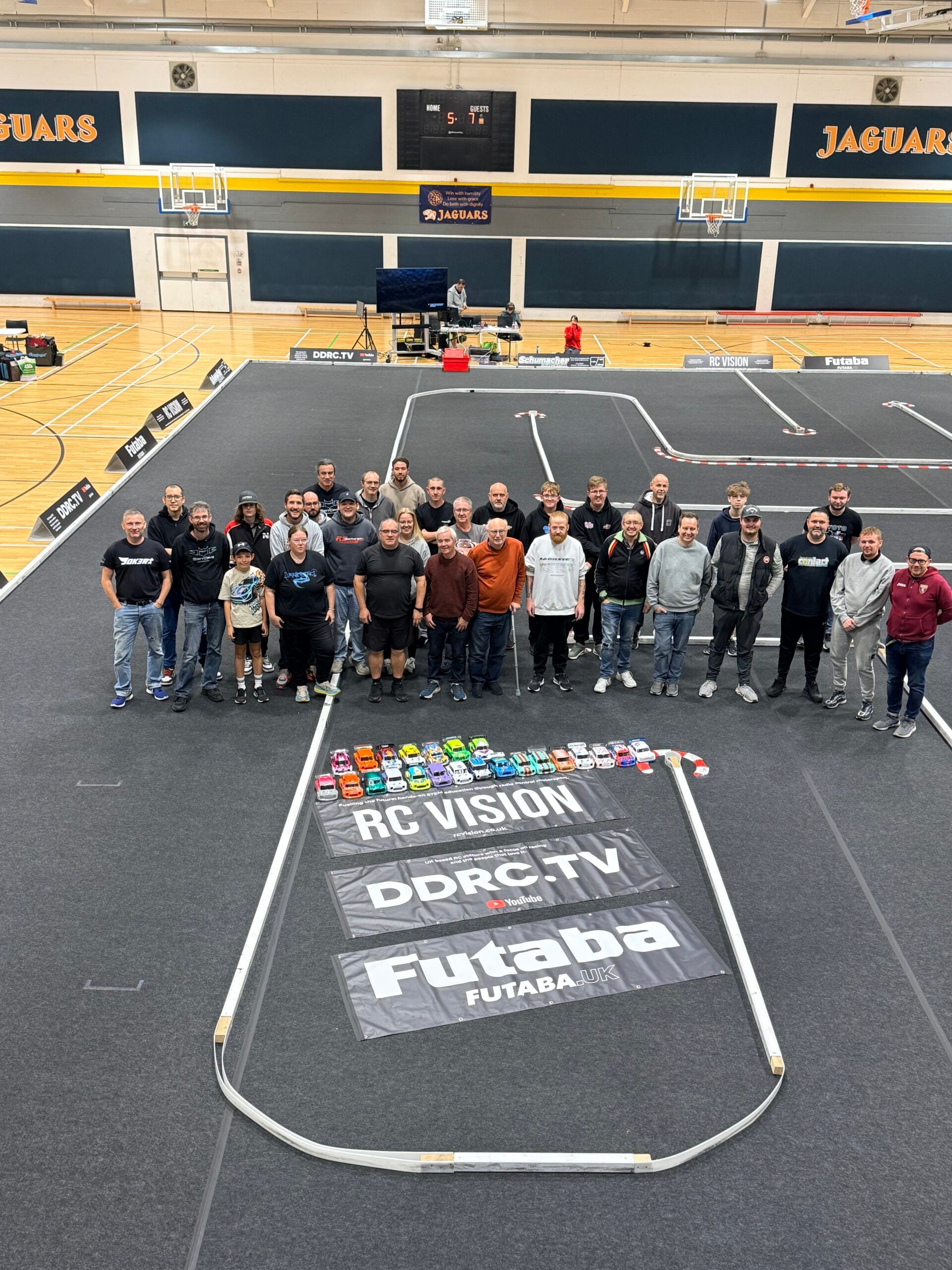 Mini Masters racers line up on the carpet track in the ACS Egham sports hall, with their Mini bodyshells and RC Vision, DDRC.TV and Futaba banners in front